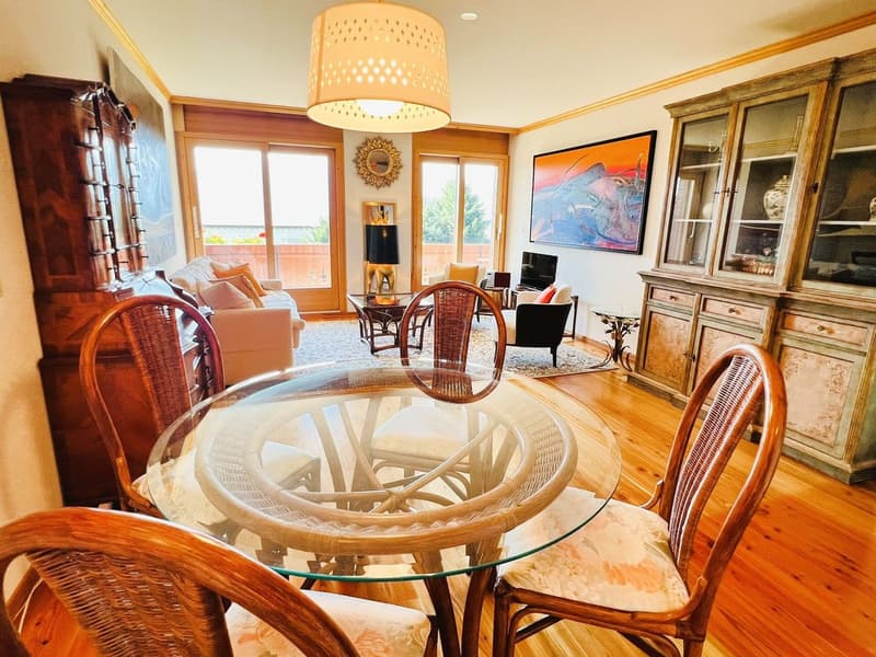 Dining area with round glass table and chairs, beige couch in living area, glass cabinets, glass windows, floor lamp, painting