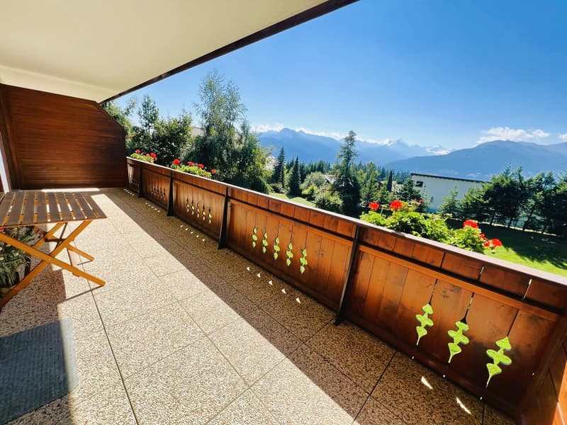 Wooden floor, wooden fence with decorative leaves, wooden table, wooden railing, flowers in pots, mountain view