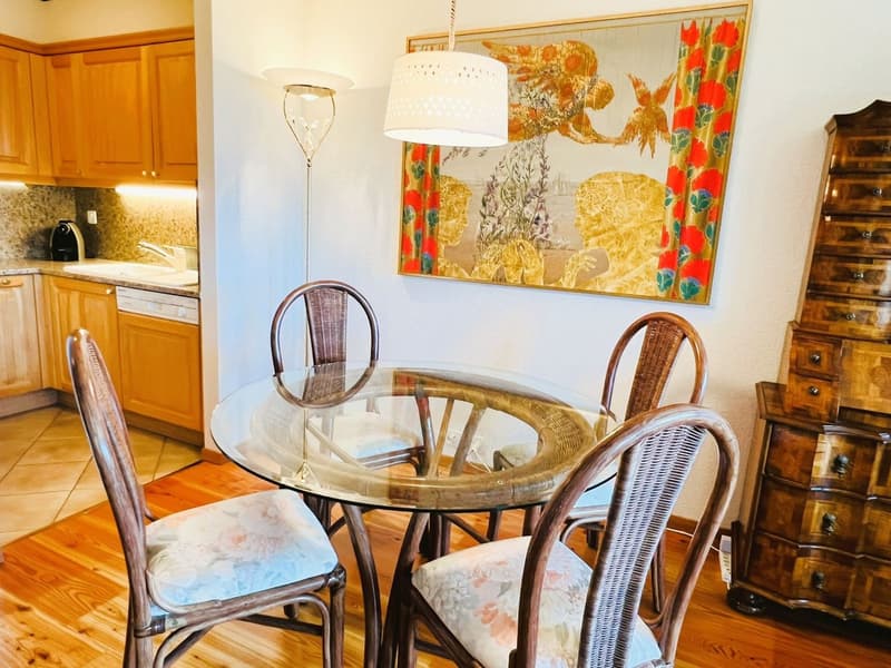 wooden floor, wooden wall cabinets, glass table, round dining chairs, hanging lamp, picture frame, kitchen area with sink, cabinet with dishwasher