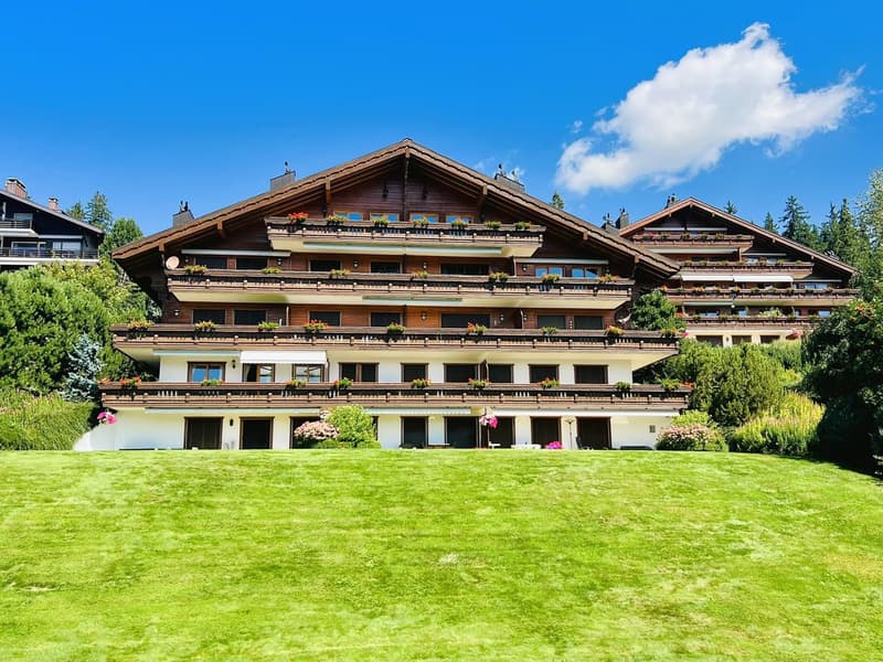 Large multi-story chalet, wooden exterior, multiple balconies with flower boxes, well-manicured lawn, surrounded by greenery