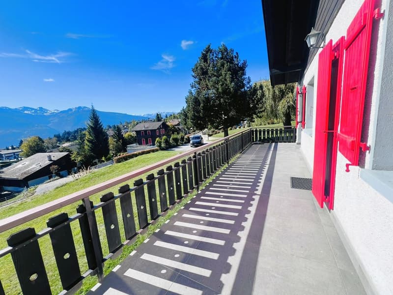 A balcony of a building with a tiled floor and black railings, overlooking a grassy field with trees and a house, mountains and a road with a car.
