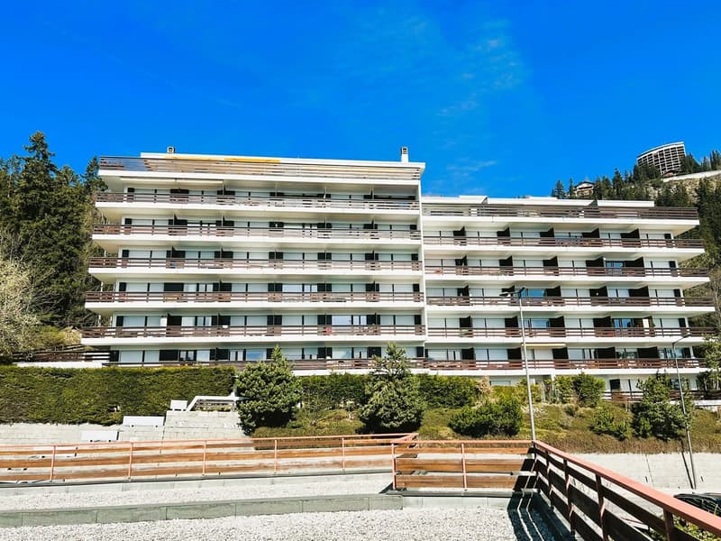 Aparthotel with white and brown facades, multiple balconies, situated on a slope with greenery and trees