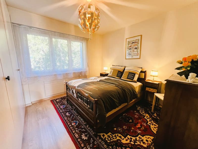 double bed, pillows, nightstands with lamps, oriental rug, white walls, large window with white curtains, picture frame, flower vase