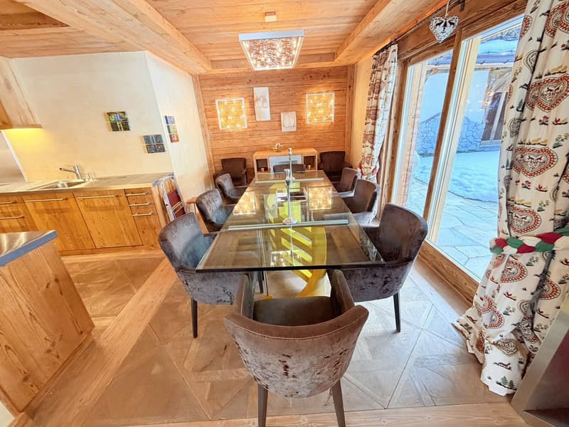 Dining area with long glass table, wooden dining chairs, wooden walls, wooden flooring, kitchen area with sink, large glass windows