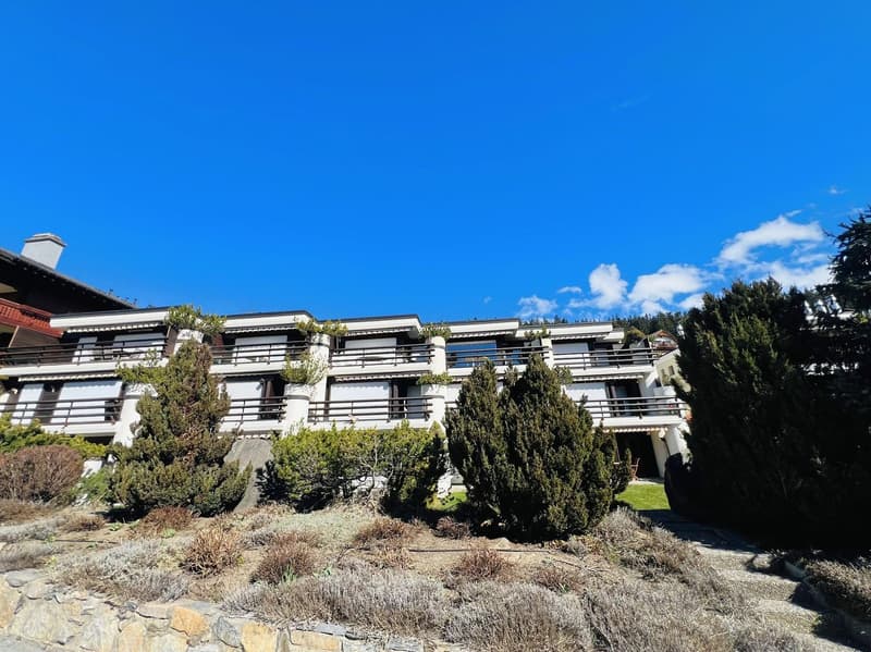 White building with multiple balconies, surrounded by trees and shrubs, stone wall at the bottom