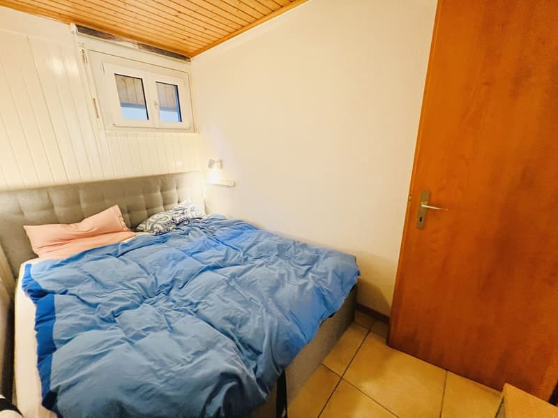 Bedroom with bed, pillows, and a lamp on the wall. Wooden ceiling and tiled floor.
