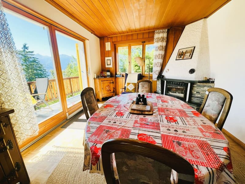 Dining room with a table and chairs, fireplace, large windows, balcony, wooden ceiling, wooden floor, rug, wooden cabinet, picture frames, and a lamp.