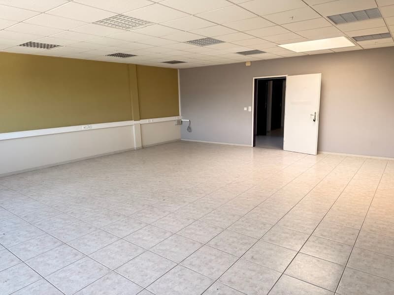 The image shows a large, open office space with a tiled floor and a suspended ceiling. The walls are painted in a neutral color scheme, and there is a white door visible in the background. The room appears to be empty, suggesting it may be a vacant office