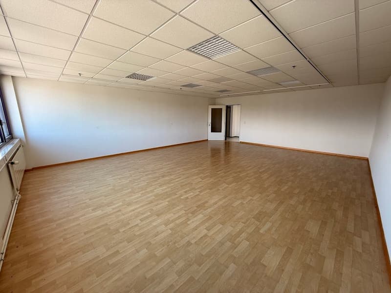 The image shows a large, empty office space with a suspended ceiling and hardwood flooring. The room appears to be a commercial or office space, with a clean and minimalist design.