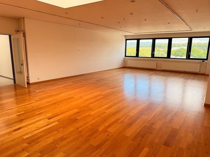 Large open room with hardwood flooring, white walls, and large windows overlooking a scenic outdoor view. The room appears to be a spacious office or commercial space with recessed lighting in the ceiling.