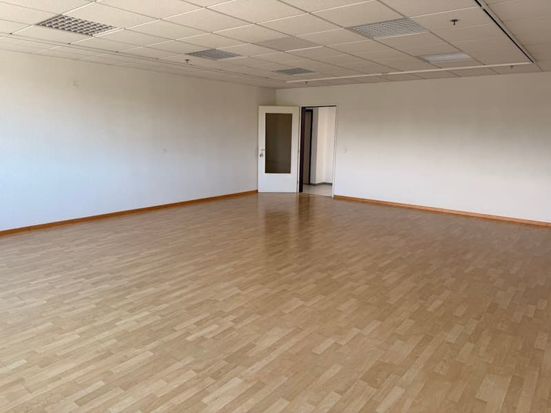 The image shows a large, empty office space with a tiled ceiling and hardwood flooring. The room has white walls and a doorway leading to another room. The space appears to be ready for use as an office or commercial space.