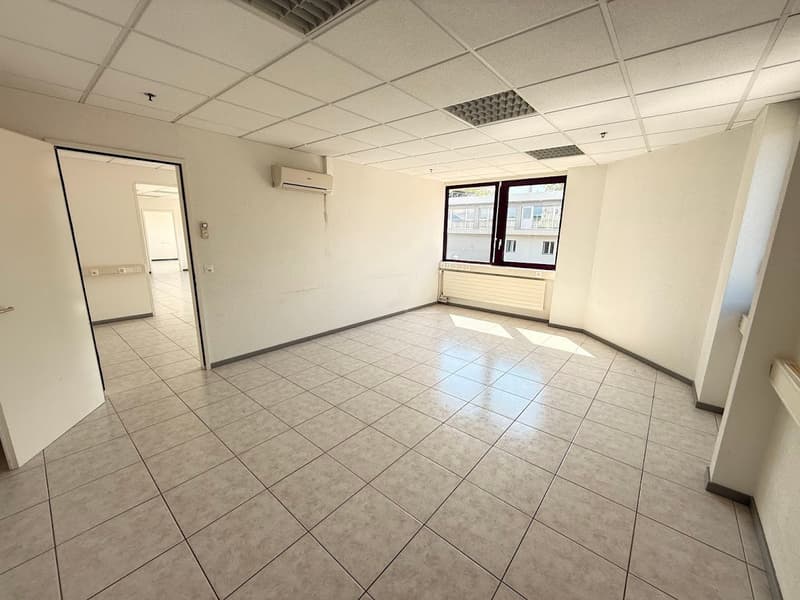 The image shows a spacious office space with a tiled floor, white walls, and a suspended ceiling with recessed lighting. The room has a large window that provides natural light. There is an air conditioning unit visible on the wall. The space appears to b