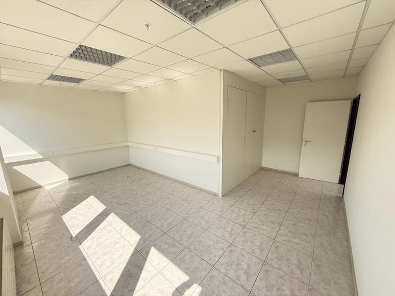 The image shows an empty office space with a tiled floor, white walls, and a suspended ceiling with lighting fixtures. The room appears to be a rectangular space with a door visible on one side.