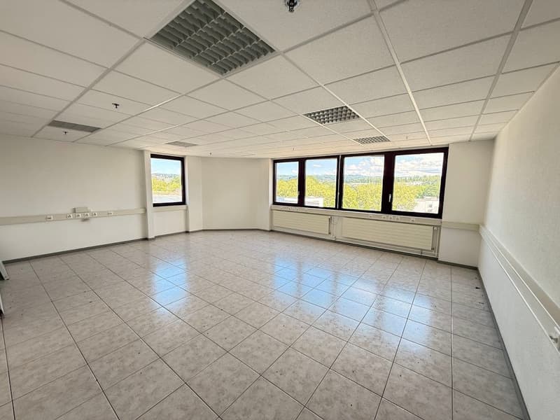 The image shows a large, open office space with a tiled floor, suspended ceiling with lighting fixtures, and large windows providing a view of the surrounding landscape. The room appears to be empty and ready for use as an office or commercial space.
