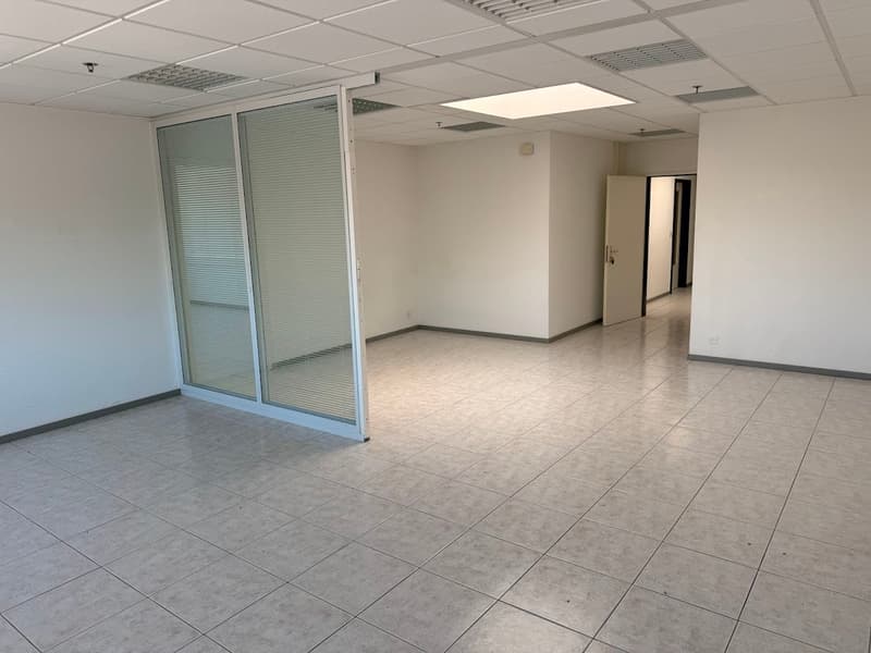 The image shows a spacious office space with a tiled floor, suspended ceiling, and glass-paneled walls. The room appears to be empty, with no furniture or other objects visible. The lighting is bright, creating a clean and professional atmosphere.