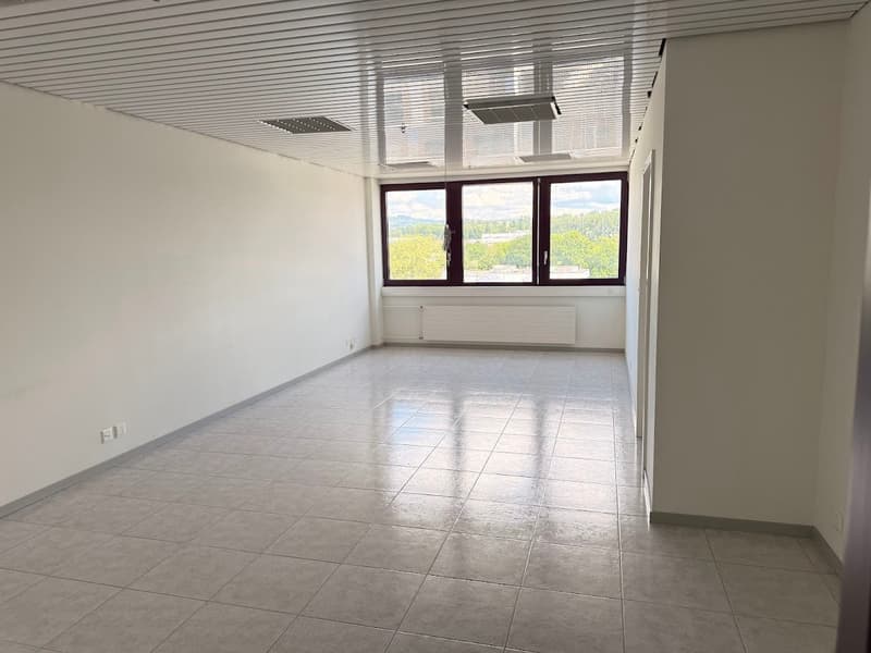 The image shows a spacious, well-lit office space with large windows overlooking a scenic outdoor view. The room has a tiled floor and a suspended ceiling with recessed lighting fixtures. The space appears to be empty and ready for use.