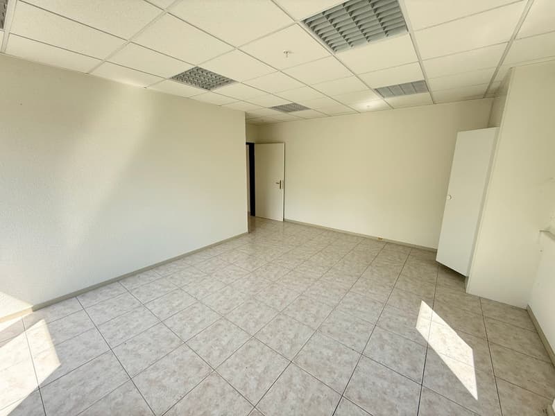The image shows an empty office space with a tiled floor, white walls, and a suspended ceiling with ventilation grilles. The room appears to be a commercial or office space, with a door visible in the background.