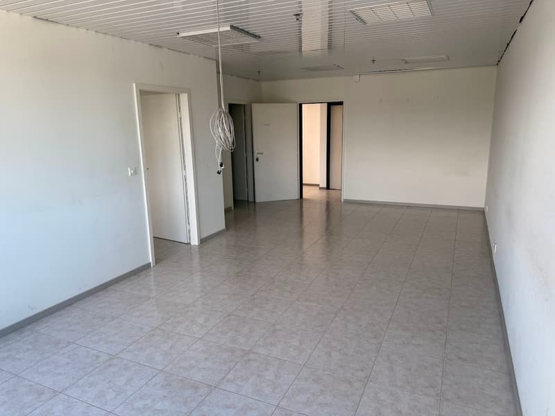 The image shows a large, open office space with a tiled floor and a suspended ceiling. The room has white walls and several doors leading to other areas. The space appears to be empty and ready for use.