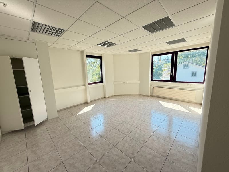 The image shows a large, open office space with a tiled floor, white walls, and a suspended ceiling with recessed lighting. The room has several large windows that provide natural light and views of the surrounding area. The space appears to be empty and