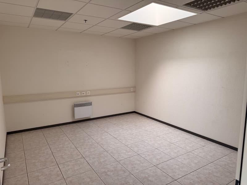 The image shows an empty office space with a tiled floor, white walls, and a ceiling with recessed lighting. There is a wall-mounted heater/air conditioning unit visible in the corner of the room.