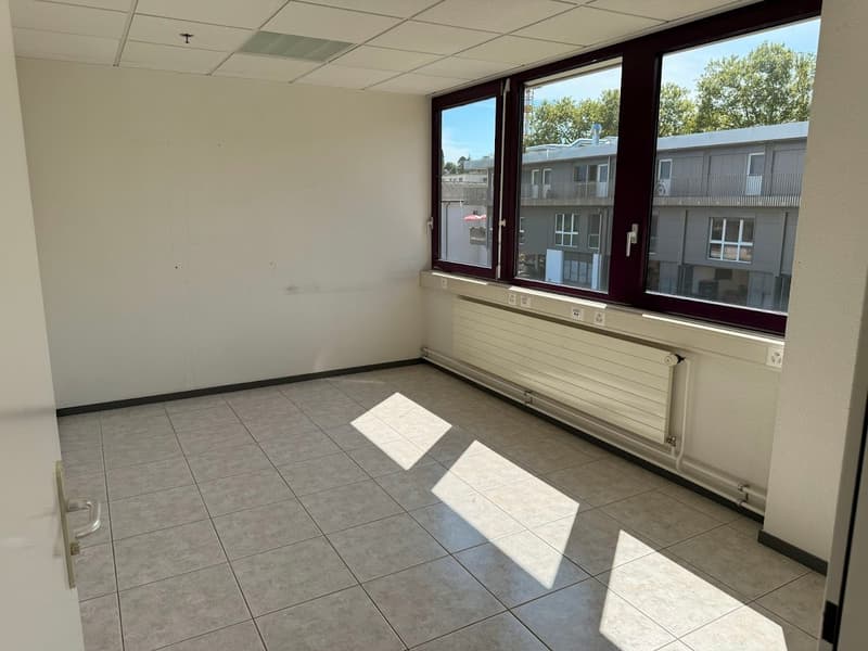 The image shows an empty office space with a tiled floor, white walls, and large windows overlooking the exterior of the building. The room appears to be a commercial or office space, with a clean and minimalist design.