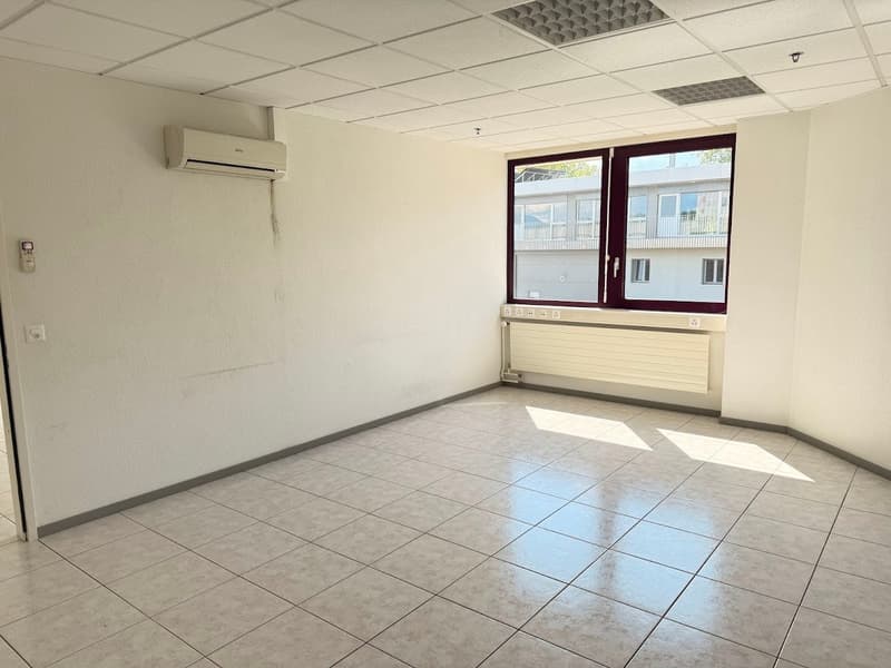 The image shows an empty office space with white walls, a tiled floor, and a large window. The room has a suspended ceiling with recessed lighting and an air conditioning unit installed. The space appears to be a commercial or office setting, ready for us