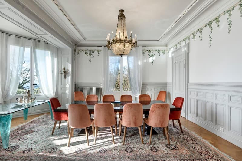 Grand dining area with chandelier, wooden chairs, large dining table, carpet, white walls, and decorative window curtains