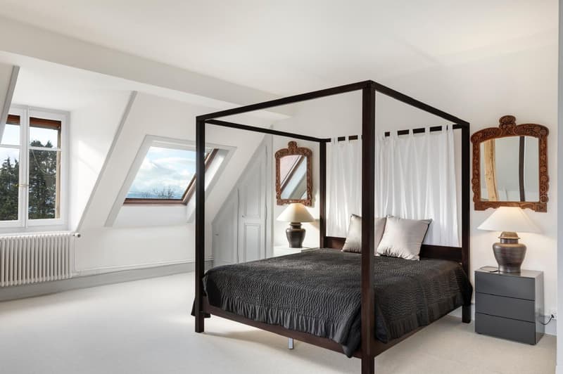 A spacious bedroom with a four-poster bed, two nightstands, lamps, and a wall-mounted mirror. Skylight windows with views of trees and mountains.