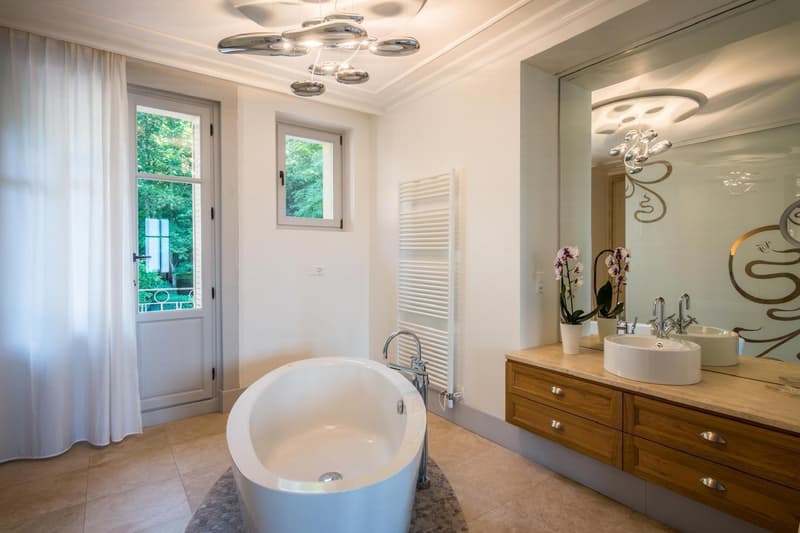 Large white bathtub, modern white sinks, wooden vanity, round mirrors, tiled floor, large windows, white curtains, radiator