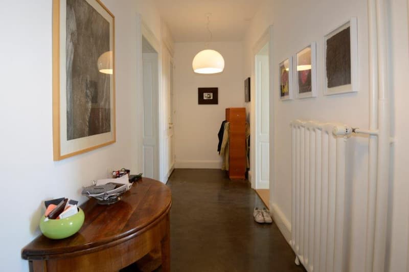 Long hallway with a wooden table and shoe rack, framed pictures on wall, lit hanging lamps, radiator
