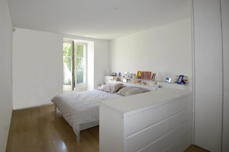 Bed with white sheet, 2 pillows, white wardrobe, shelf above the bed with books and picture frames, wooden floor, white walls, glass door.