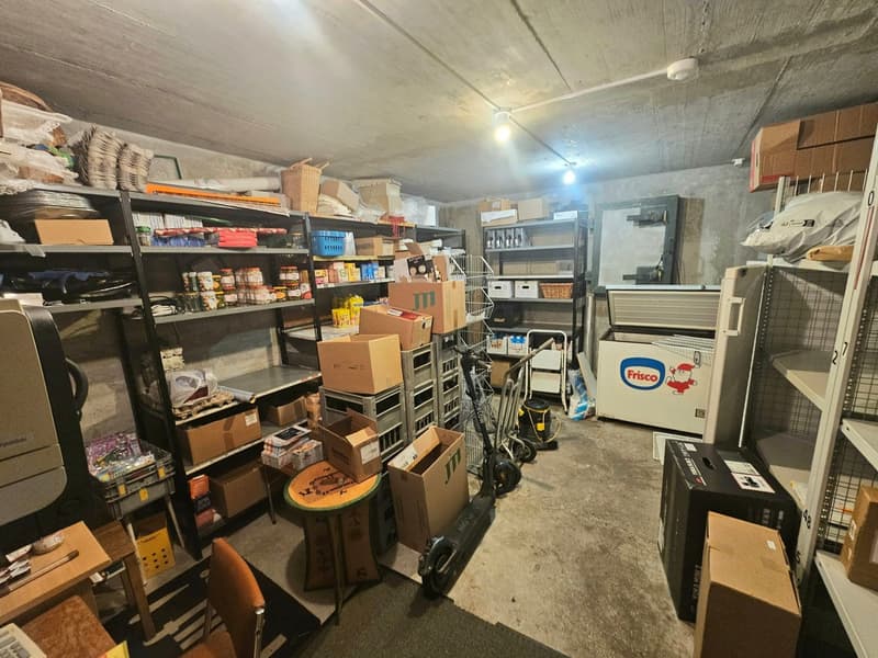 A storage room filled with various boxes, shelves, and items. There are baskets, jars, bottles, cans, and other miscellaneous items on the shelves. A scooter, a chair, and a table are also present. A refrigerator and a trolley are in the room, along with