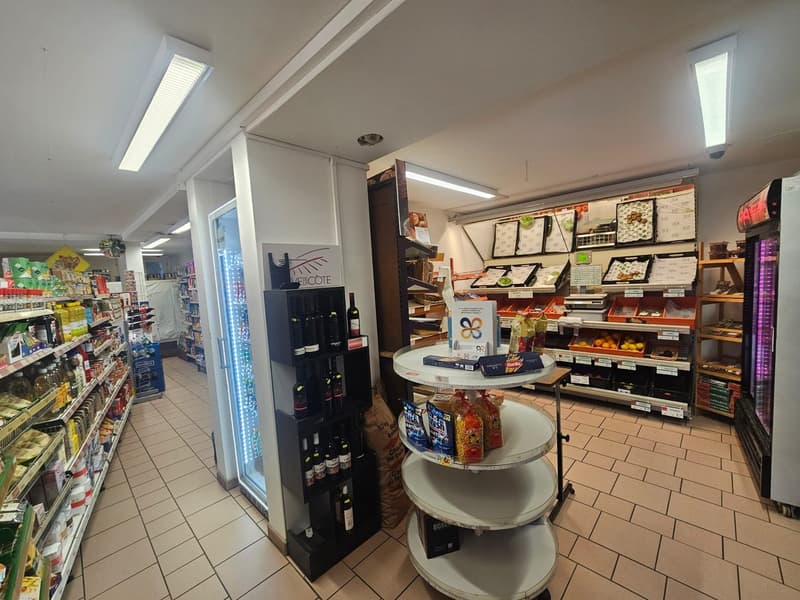 Interior of a store with various products on display, including bottles, food items, and boxes. A refrigerator and a display case are visible, along with ceiling lights and a ceiling fan.