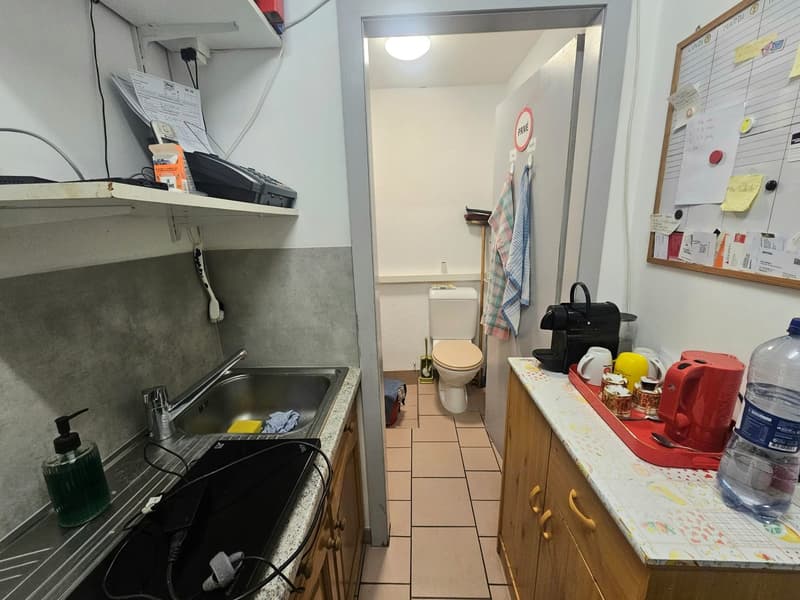 Small kitchen with sink, faucet, soap dispenser, and dishwasher. A toilet and toilet paper are visible in the adjacent room. A kettle, cups, and a water bottle are on the counter. A board with papers and a box is on the wall.