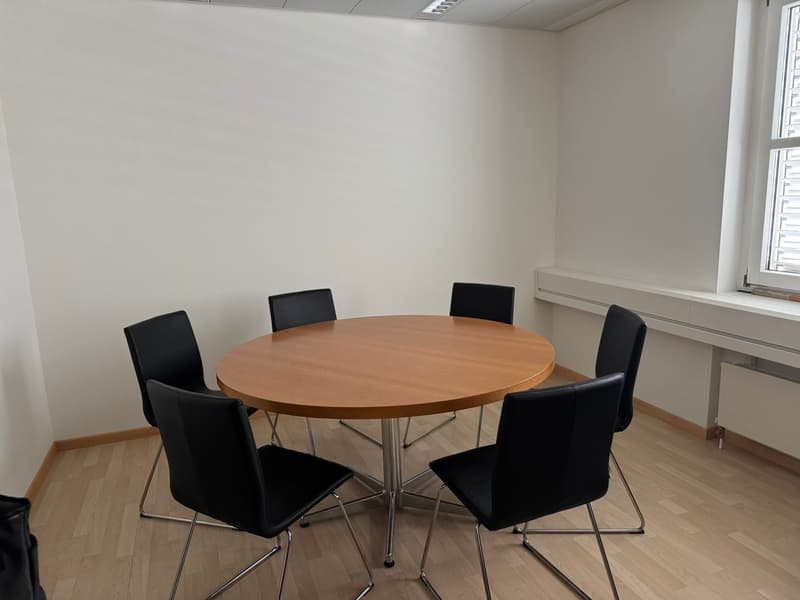 Meeting room: round wooden table, six black chairs, wooden floor, white walls, radiator, window.