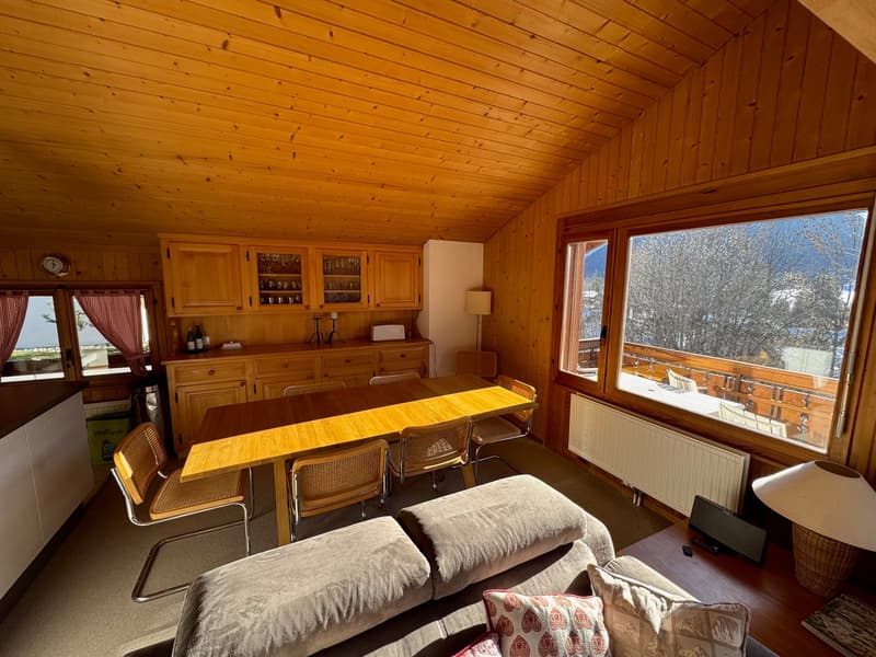 Open plan living room and dining area, large windows, wooden ceiling and walls, radiator, table and chairs, sofa, coffee table, lamps