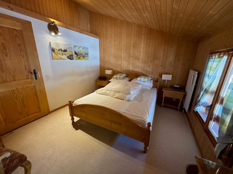 A bedroom with wooden walls and floor, bed, pillows, nightstands, lamps, radiator, wooden door, chair, paintings, and curtains.