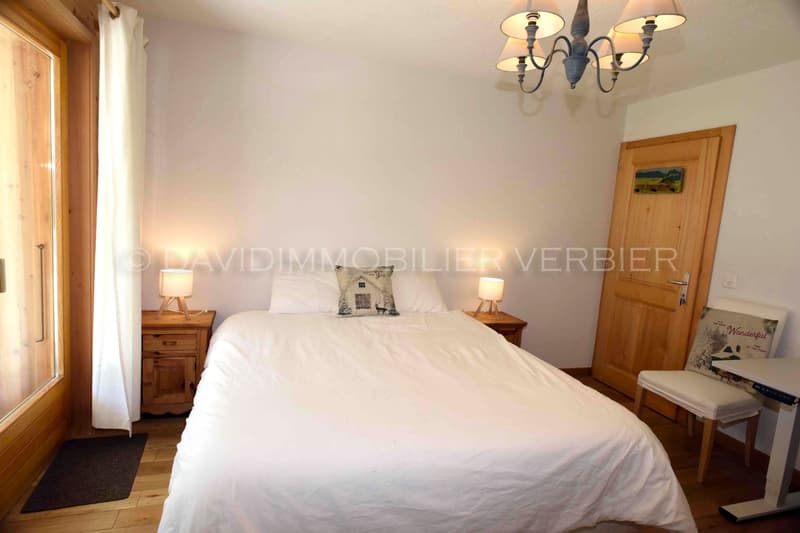 White painted walls, wooden floor, bed with white sheets and pillows, bedside tables with lamps, window with curtain, wooden door, chair with cushion, desk, chandelier