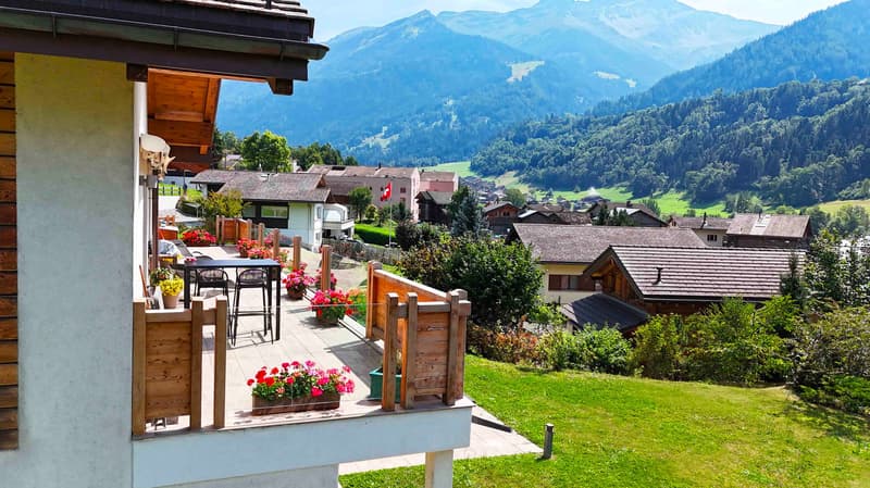 Apartment with balcony, mountain view, wooden railing, outdoor seating with table and chairs, flower pots, green lawn, several buildings and a mountain range in the background.