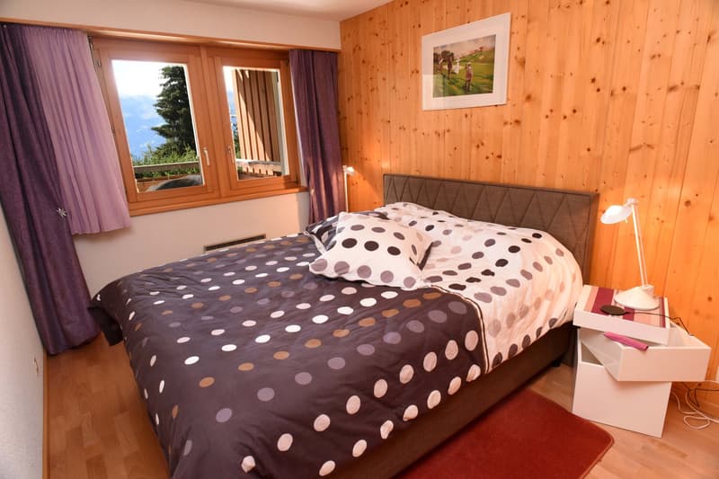 Bedroom with wooden walls, bed with polka-dot bedspread, lamp on nightstand, framed picture, window with curtains, small rug, and a view of the mountains outside.