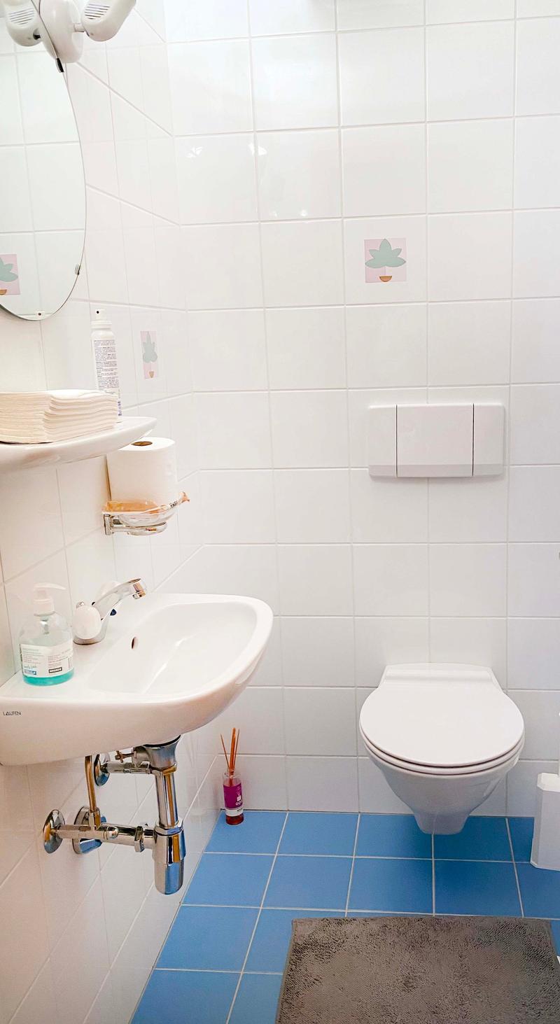 Small bathroom with white tiles, blue floor tiles, sink with soap and hand towel, toilet, round mirror, toilet paper