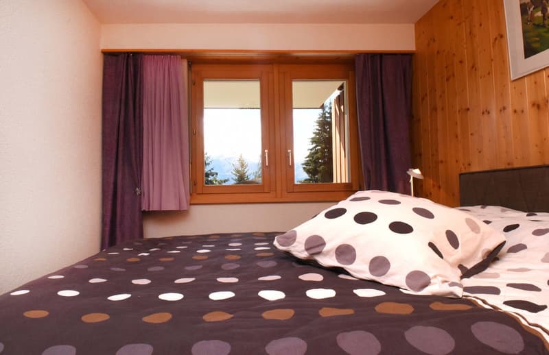 Wooden walls, window with view to the mountain, double bed with polka dot bedding, bedside table with lamp, framed picture