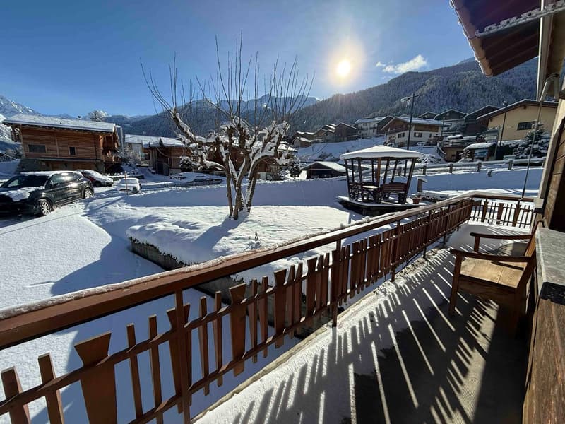 balcony with wooden railings, wooden chair, snow-covered ground, snowy mountains, wooden house, gazebo, parked cars