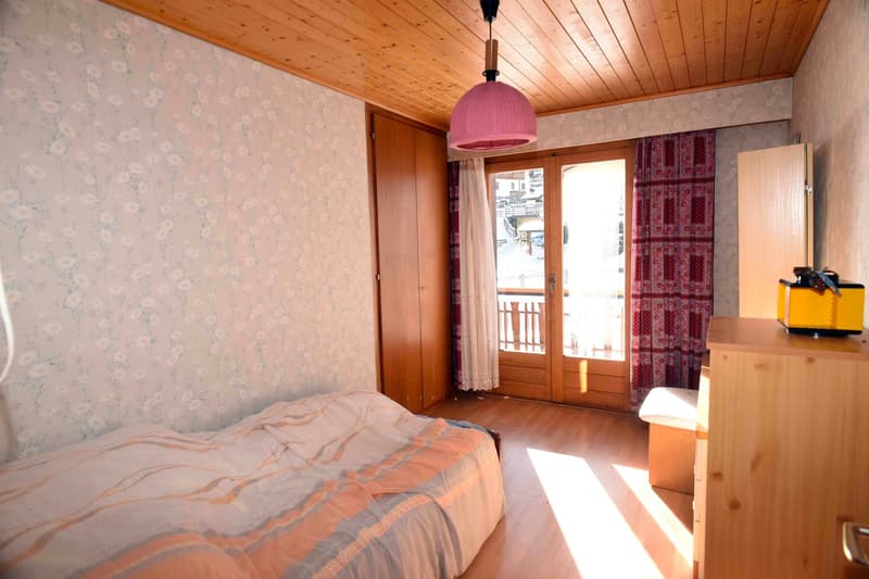 bedroom with a bed, wooden wardrobe, desk with a small bench, floral wallpaper, pink lamp, double sliding door with a balcony view