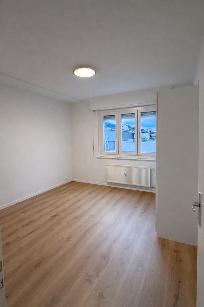 large empty room, wooden floor, white walls, large window, ceiling light, radiator