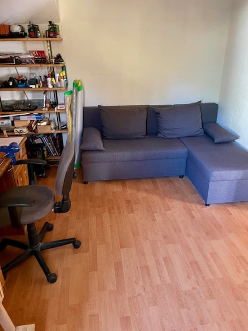 A small room with a blue couch, a wooden desk, a chair, and a shelf with books and other objects.