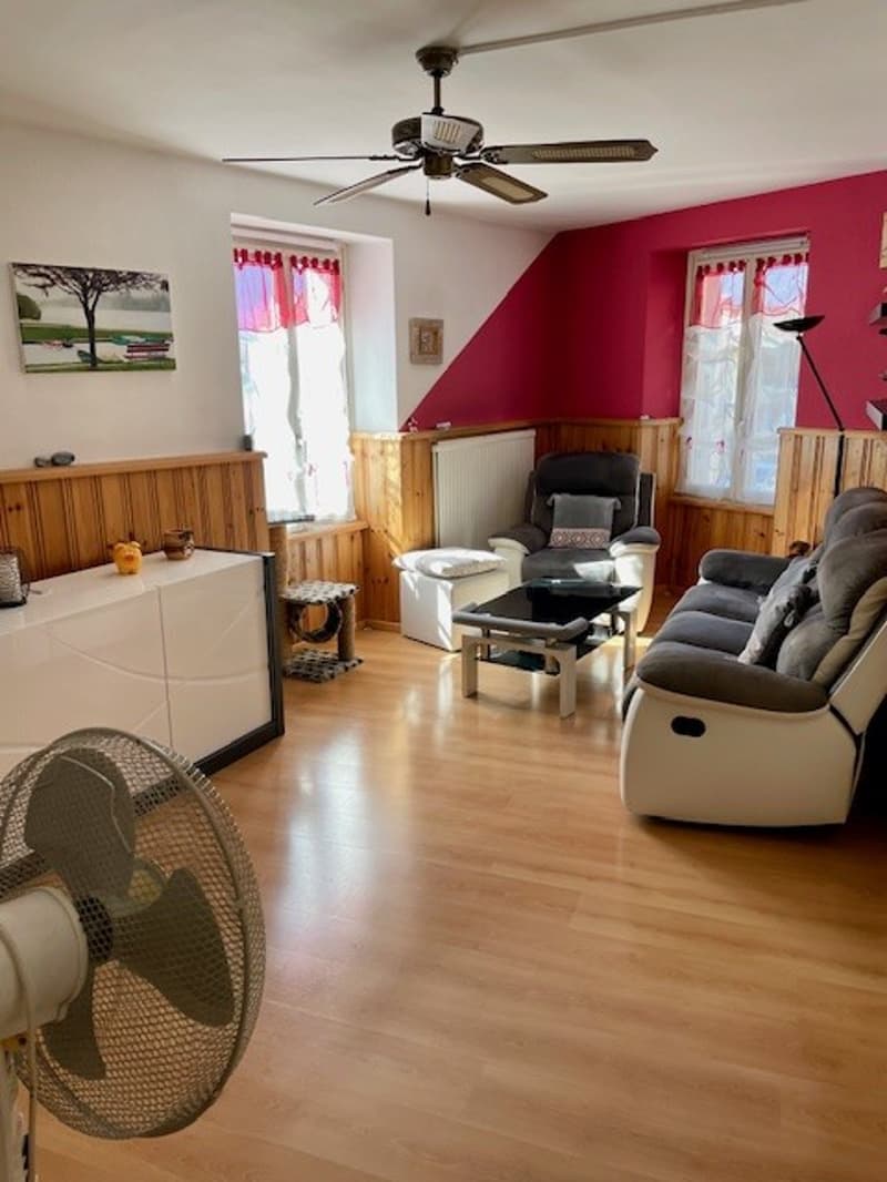 living room with sofa, two armchairs, coffee table, fan, ceiling fan, wooden floor, wooden wall paneling, radiator, paintings, red accent wall