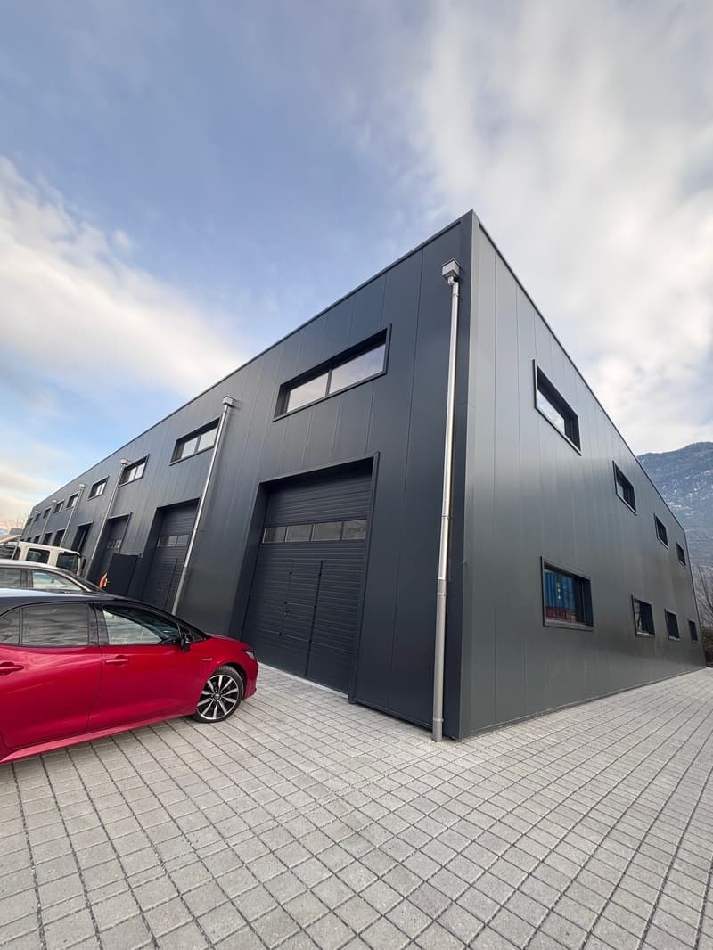 Modern industrial building, dark grey exterior, multiple windows, double garage doors, surrounded by gravel parking area