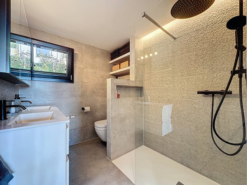Modern bathroom with walk-in shower, toilet, double sink, built-in shelves, and window with trees view
