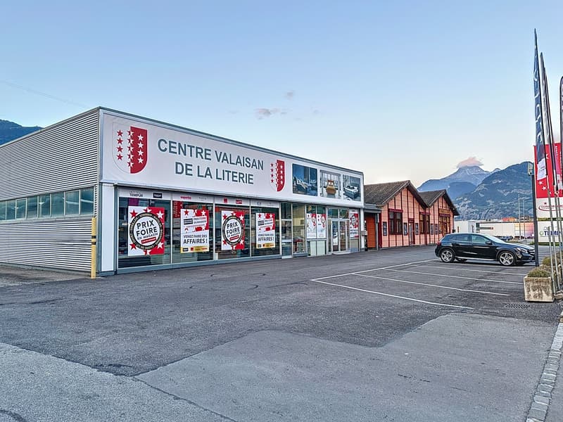 large building, sign reading 'CENTRE VALAISAN DE LA LITERRIE', red roof, parking area, mountains in the background
